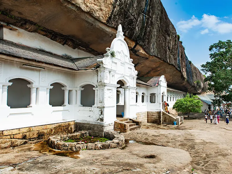 The white colonial-style arched entrance facade of the Dambulla Cave Temple built directly beneath the massive overhanging rock, a major UNESCO site near Sigiriya
