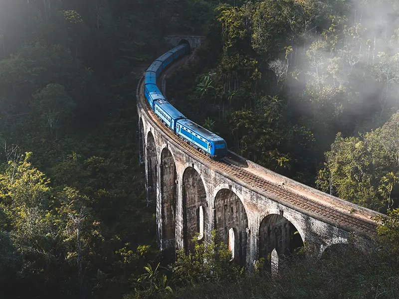 The iconic Nine Arch Bridge with a blue train crossing it, showcasing the incredibly scenic railway as a primary way of getting around Sri Lanka.