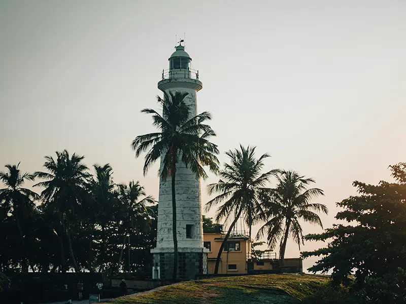 Close-up of the white Galle Fort Lighthouse surrounded by palm trees at sunset, a popular sight among things to do in Galle Fort.