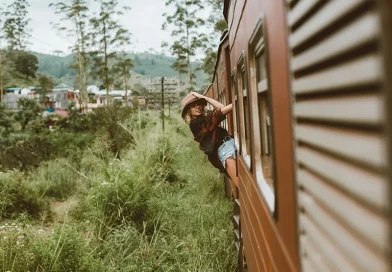 A traveler in a hat leans out of the window of a red carriage, enjoying the scenic views on the famous Kandy to Ella train journey.