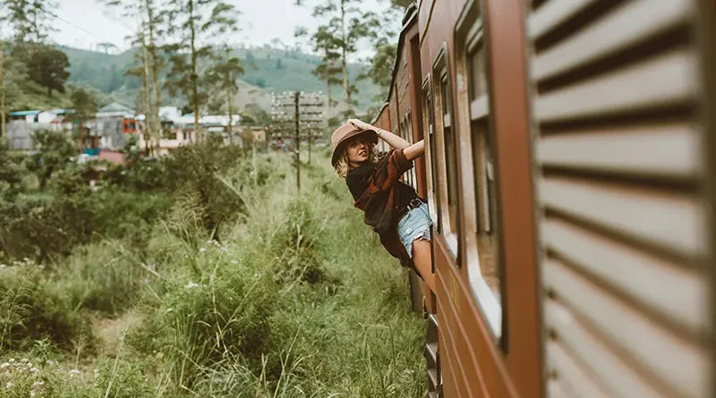 A traveler in a hat leans out of the window of a red carriage, enjoying the scenic views on the famous Kandy to Ella train journey.