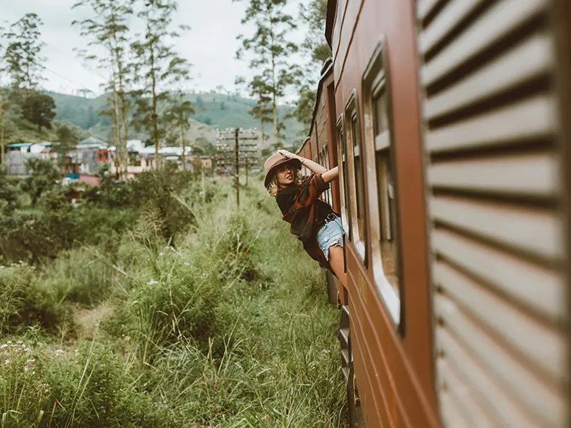 A traveler in a hat leans out of the window of a red carriage, enjoying the scenic views on the famous Kandy to Ella train journey.