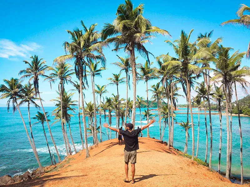 Things to do in Mirissa Sri Lanka: Traveler with arms wide open at the iconic Coconut Tree Hill viewpoint overlooking the turquoise ocean.