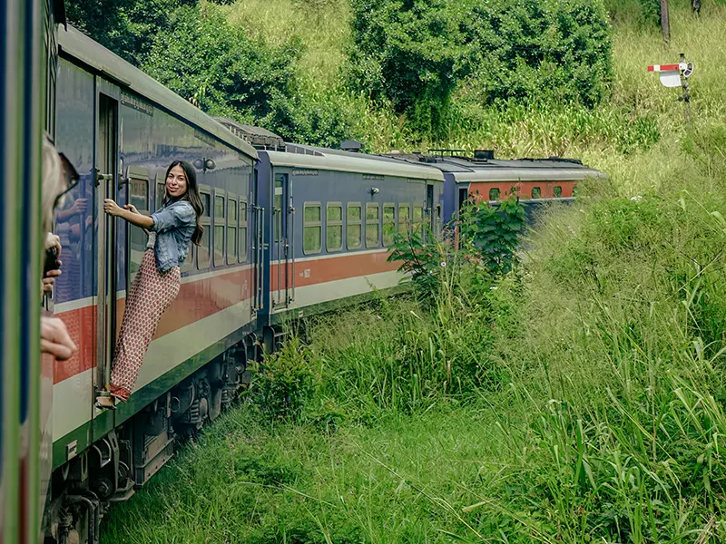 A smiling female traveler leaning out of the open doorway of a carriage, enjoying the adventurous Kandy to Ella train ride through the overgrown countryside.