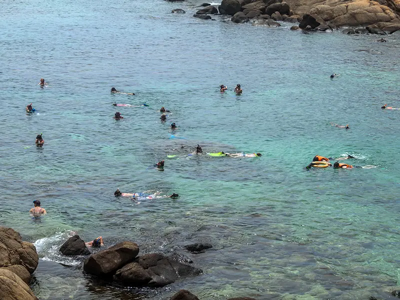 Tourists snorkeling in the shallow, clear turquoise waters of Pigeon Island National Park near Trincomalee, surrounded by rocky outcrops and a coral reef, famous for blacktip reef sharks and sea turtles.