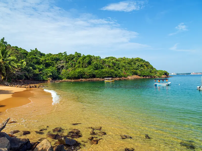 A secluded, sandy cove known as Jungle Beach Unawatuna, featuring calm, clear turquoise water, dense surrounding foliage, and anchored boats, popular for swimming and snorkeling.