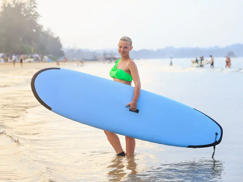 A smiling woman holding a light blue surfboard in the shallow water of Weligama Beach, representing the popular beginner surf lesson activity and a top things to do in Weligama highlight.